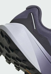 Zapatilla de correr con un upper de malla púrpura y suela de goma negra con tacos pronunciados; detalles en gris claro en el talón y los lados.