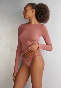 Langsleeve, getailleerde top van zachtroze stof, gecombineerd met een bijpassende thong met kantaccenten. Gladde textuur, subtiel ontwerp en verfijnde kantdetails.
