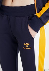 Personne portant un pantalon de sport bleu marine et jaune, une main dans la poche et un logo hummel visible sur la jambe.