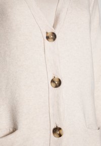 Světle béžový cardigan s výstřihem do V, se čtyřmi tmavě hnědými knoflíky a dvěma předními kapsami. Měkká textura a volný střih.