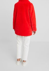 Abrigo rojo de piel sintética con textura suave, mangas largas y cuello con solapa, combinado con jeans blancos y zapatillas.