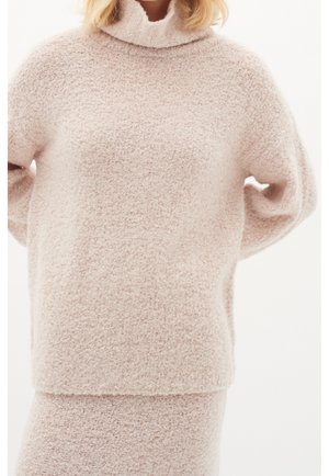 Kobieta ubrana w jasnoróżowy, teksturowany, długi sweter z golfem i dopasowaną spódnicę, stojąca na tle jednolitego białego tła.