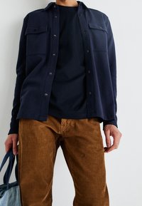 Námořnická košile na knoflíky s dvěma náprsními kapsami, přes tmavé tričko, v kombinaci s hnědými kordáčky, držící modrou tašku.