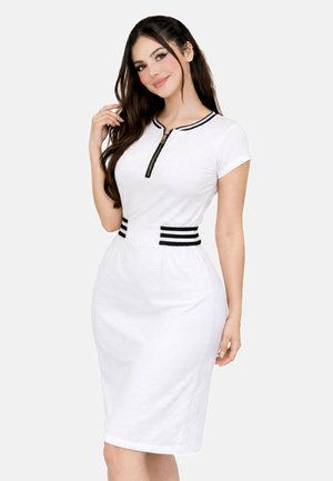 Młoda kobieta z długimi ciemnymi włosami, ubrana w dopasowaną białą sukienkę z czarnym paskowanym zamkiem błyskawicznym na kołnierzu i w pasie, pozująca na jednolitym tle.