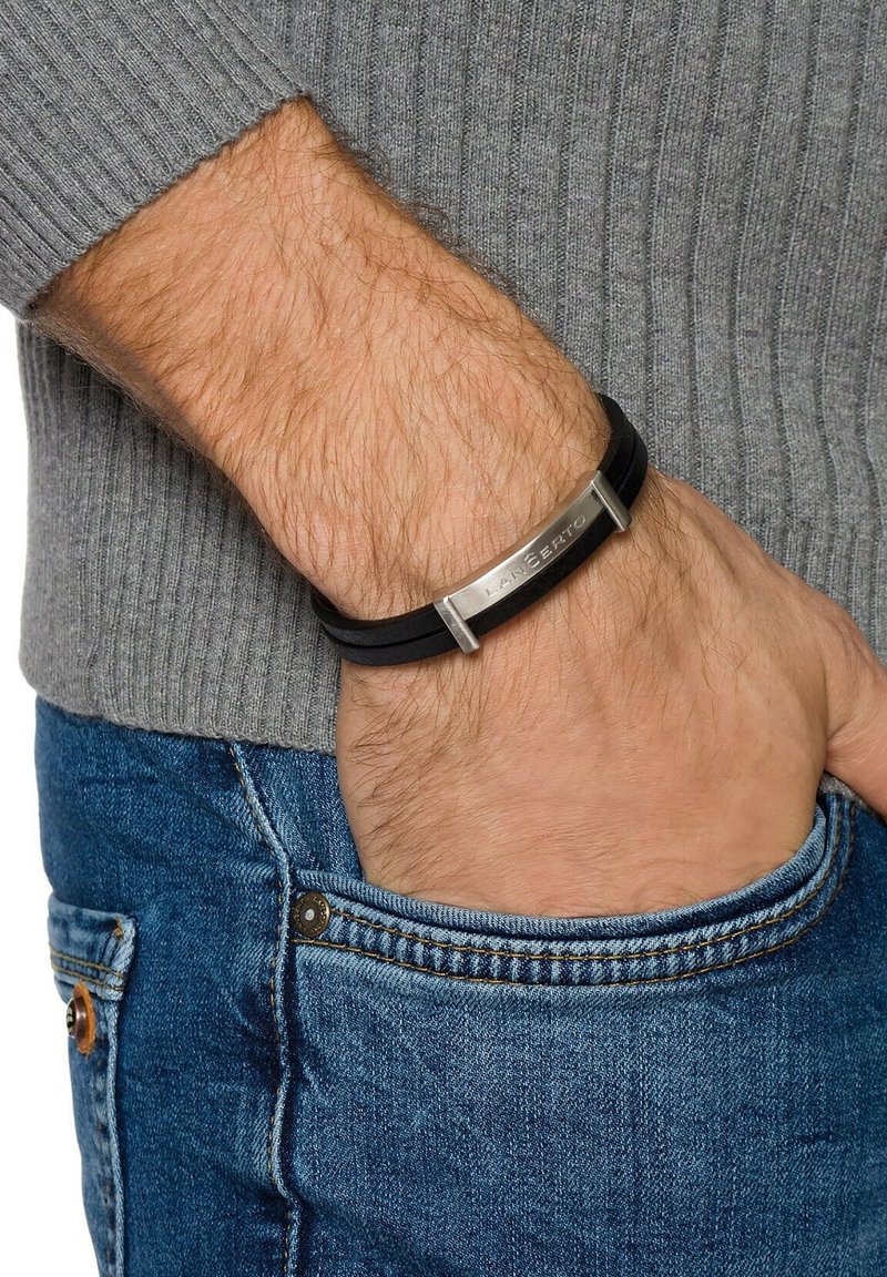 Main d’homme dans une poche en denim bleu portant un pull gris et un bracelet noir avec une plaque métallique gravée "LANCERTO" au poignet.