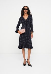 Vrouw in een marineblauwe jurk met stippen, zwarte hoge hakken, donkere zonnebril, met een lichtroze clutch tas in haar hand, staand tegen een witte achtergrond.