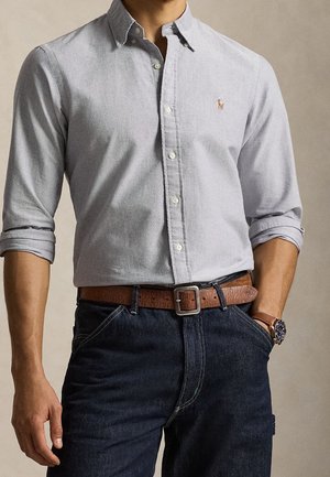 Mand iført en lysegrå skjorte med knapper og opkrogede ærmer, mørke jeans, brun læderbælte og et armbåndsur med brun rem.
