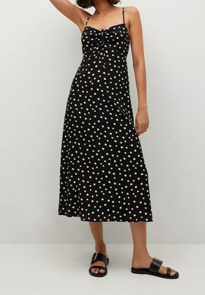 Robe midi noire à pois blancs, bretelles spaghetti, détail froncé sur le buste, et tissu fluide. Associée à des sandales noires.