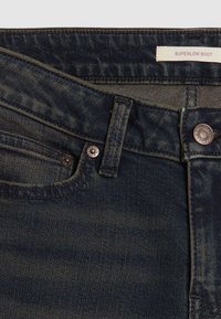 Jean en denim bleu foncé avec une coupe super basse style bootcut. Comprend des éléments en cuivre et des variations subtiles de texture.