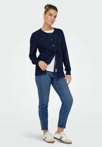 Mornarska pletena kardigan s uzorkom kablova, zatvaranjem na gumbe i detaljem s vezanjem u struku, uparen s plavim trapericama i bijelim tenisicama.