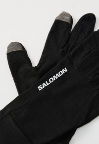 Gants noirs en matériau doux, dotés d'une pointe texturée grise sur le pouce et l'index, avec un logo "SALOMON" blanc sur le poignet.