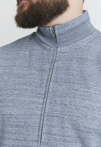 Gai pelēks zip-up džekets ar augstu apkakli, ar teksturētu audumu un smalku heather rakstu. Frontes daļā redzama rāvējslēdzēja detaļa.