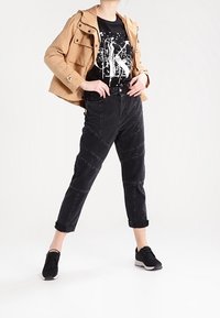 Chaqueta ligera color beige, camiseta gráfica negra, pantalones ajustados negros con costuras y zapatillas negras, exhibidos sobre un fondo blanco.