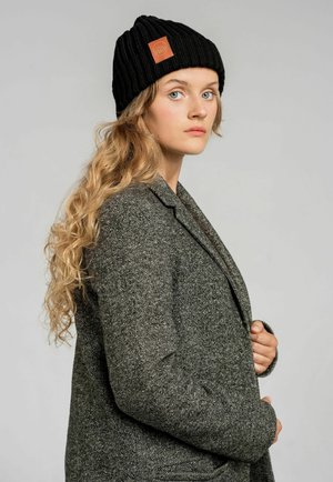 Czarna, prążkowana czapka z daszkiem z skórzaną naszywką, noszona z szarym, wełnianym żakietem w jodełkę. Model ma falujące blond włosy. Neutralne tło.
