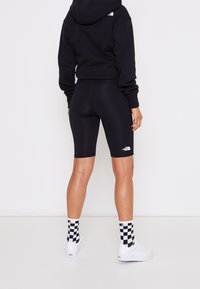 Černá krátká mikina s kapucí, černé cyklistické šortky, bílé logo, doplněné o bílé boty a kostkované ponožky. Hladká textura, přiléhavý střih.