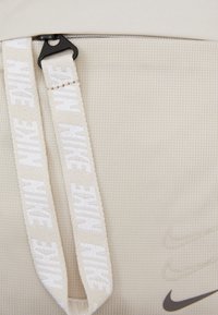 Närbild av beige tyg med strukturerat mönster, två beigefärgade Nike-logoband fästa vid en svart spänne, och tre präglade Nike swooshes.