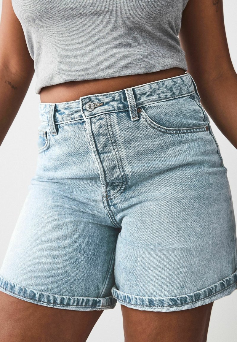 Femme portant un short en jean bleu clair taille haute et un débardeur gris côtelé court, montrant le ventre et une partie des bras.