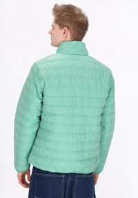 Giubbotto puffer verde menta con colletto alto, texture trapuntata e polsini elasticizzati, indossato sopra un indumento in denim blu scuro.