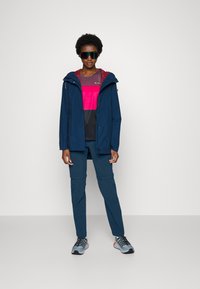 Marineblauwe waterdichte jas met een capuchon over een roze en zwarte top. Gecombineerd met blauwe broek en grijs-zwart sneakers met oranje accenten.