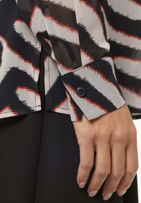 Sheer blouse featuring a abstract pattern in black, beige, and red, with a cuff detail and a round black button.