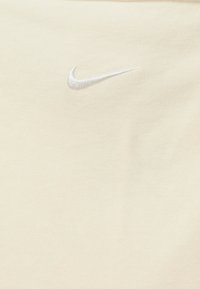 Krémové tričko vyrobené z měkké látky, na hrudi zdobené malým vyšívaným bílým logem Nike swoosh. Jednoduchý design, krátké rukávy.