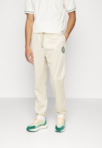 Man i krämfärgade joggerbyxor med randigt mönster och emblem, vit kortärmad skjorta, vita strumpor och gröna samt beige sneakers, som står.