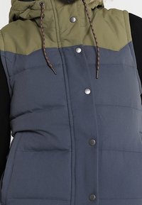 Vadderad väst med huva, med en grön övre sektion och en marinblå nedre sektion. Fäst med silverfärgade tryckknappar, med horisontell sömnad.