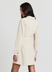 Beiges Cordkleid mit Rückenpasse, langen Ärmeln und geknöpften Manschetten. Hat eine taillierte Passform mit Gürtel und eine gerippte Textur.