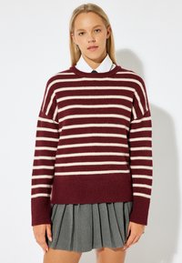 Maglione a righe bordeaux e crema con vestibilità comoda, orlo a costine e scollo rotondo, abbinato a una gonna grigia plissettata.