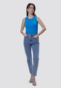 Top sin mangas azul con acentos de volantes en las costuras laterales, combinado con unos jeans de mezclilla clara que presentan rayas blancas verticales y un corte ajustado, usados con sandalias azules.