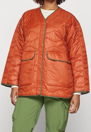 Femme portant une veste matelassée orange rouille avec une bordure verte et deux grandes poches avant, associée à un pantalon cargo vert.
