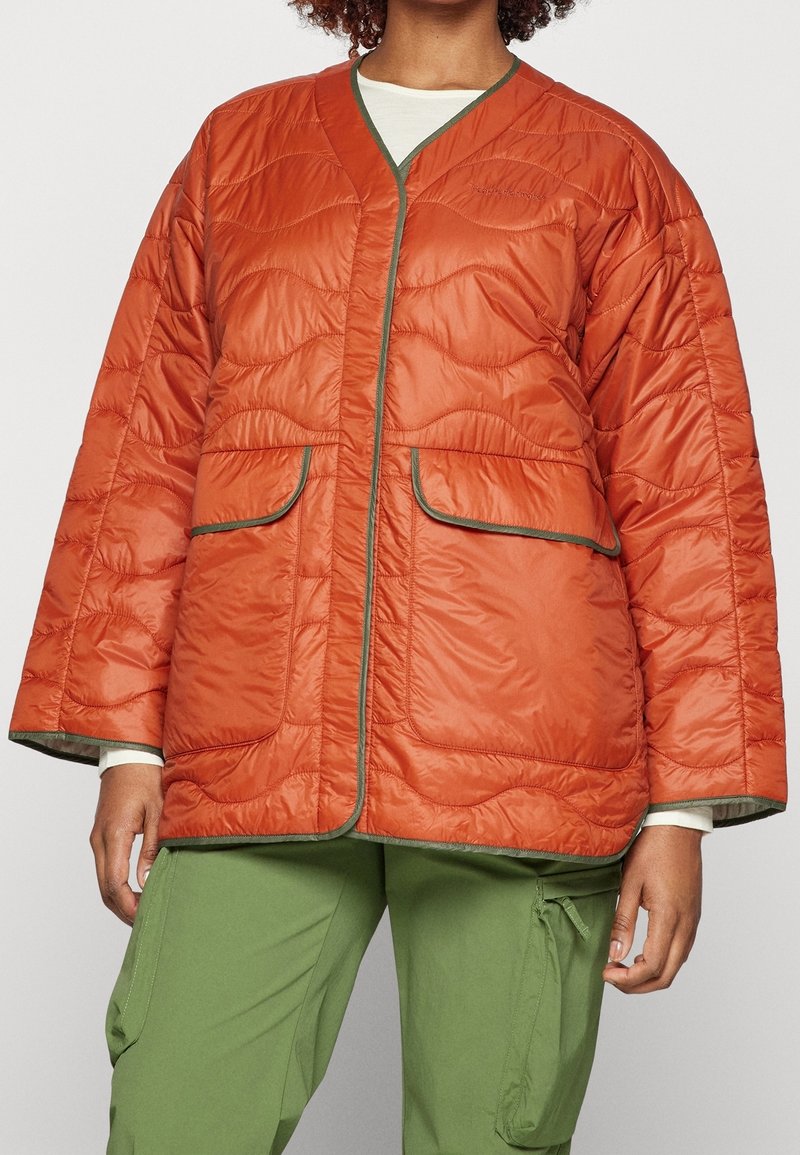 Femme portant une veste matelassée orange rouille avec une bordure verte et deux grandes poches avant, associée à un pantalon cargo vert.