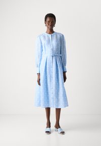 Light blue, long-sleeved dress with white polka dots, button front, belted waist, and pleated skirt. Paired with light blue slip-on shoes.