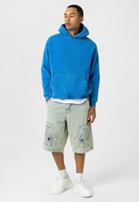 Blauwe hoodie met een voorzak; gecombineerd met lichte denim shorts met bloemenborduurwerk en versleten details; witte sneakers.