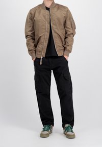 Persona che indossa una giacca bomber marrone, pantaloni cargo neri e sneaker beige con lacci verdi, in piedi con le mani nelle tasche.