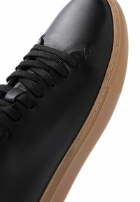 Sneaker in pelle nera con finitura testurizzata, caratterizzato da lacci neri, una punta liscia e una suola in gomma color caramello. Accenti cuciti vicino ai passanti.