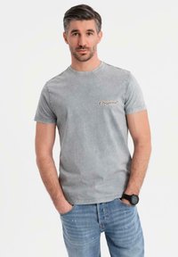 Camiseta de algodón gris con cuello redondo y mangas cortas, que presenta el logo "Original" en marrón en el pecho. Combinada con vaqueros azules.