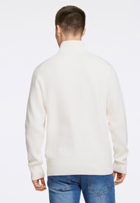 Hvid ribstrikket sweater med høj krave og lange ærmer, parret med blå denim jeans. Enkelts design uden bemærkelsesværdige mønstre eller accenter.