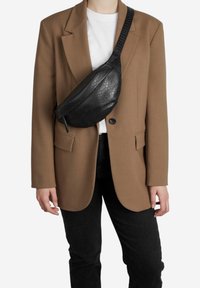 Melnas mākslīgās čūsku ādas jostas somiņas, ar noapaļotu formu, ar rāvējslēdzēju un regulējamu jostu, valkā virs baltā T-krekla un brūnā žaketes.