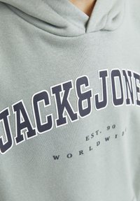 Felpa grigia con logo frontale che riporta "JACK & JONES" in stampa blu navy in grassetto. Presenta un cappuccio con cordino e una texture morbida.