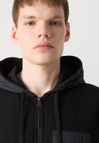 Νέος άνδρας με ανοιχτόχρωμο δέρμα και μπλε μάτια, φορώντας μια μαύρη φούτερ με φερμουάρ και επενδεδυμένη κουκούλα μπροστά σε λευκό φόντο.