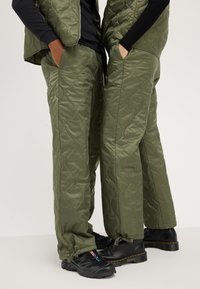 Gilet et pantalon olive vert matelassés avec une texture brillante, comportant des poches amovibles et des poignets ajustables. Chaussures noires avec des détails de logo.