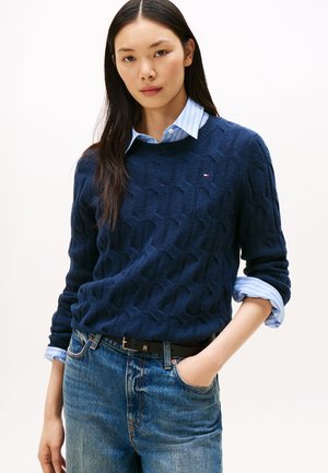 Giovane donna con capelli lunghi e scuri che indossa un maglione lavorato a trecce blu navy sopra una camicia a righe azzurre e jeans blu, mano in tasca.