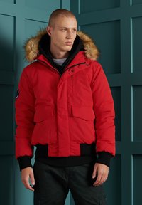 Rød polstret jakke med en sort, ribbet kant og manchetter. Har en hætte med pelsforing og flere frontlommer. Mat finish, varm design.