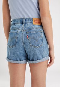 Shorts de mezclilla con un lavado azul claro, con puños enrollados, bolsillos traseros con costuras amarillas y una etiqueta de cuero marrón con el logo de la marca.