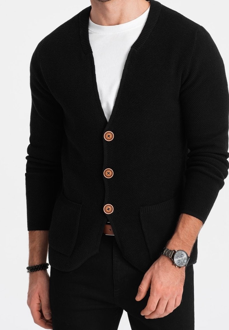 Homme portant un cardigan noir texturé avec trois boutons marron, un t-shirt blanc, un pantalon noir, une montre au poignet gauche et un bracelet noir au poignet droit.