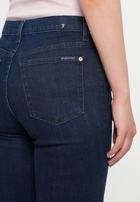 Jeans negri închisi, cu un design ajustat, având o buzunar posterior cu un eticheta cusută și un mic buton metalic la talie.