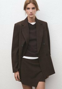 Blazer marrone a quadri con doppi bottoni, abbinato a un maglione scuro e una minigonna, sovrapposti a una camicia bianca. Tessuto strutturato.