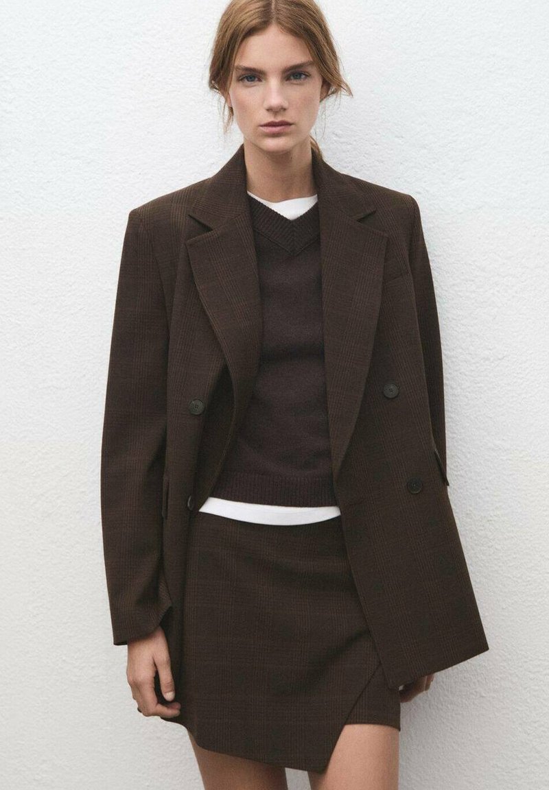 Blazer marrone a quadri con doppi bottoni, abbinato a un maglione scuro e una minigonna, sovrapposti a una camicia bianca. Tessuto strutturato.