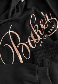 Černá souprava s kapucí a tepláky s velkým kovovým textem v barvě růžového zlata, hladkým bavlněným materiálem a elastickým pasem.
